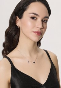 Srebrny naszyjnik z niebieskim kamieniem i małym dekoracyjnym zawieszeniem, noszony na czarnym satynowym topie z cienkimi ramiączkami. Gładka tekstura i prosty design.