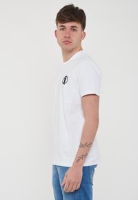T-shirt bianco a maniche corte in cotone, con un piccolo logo nero sul lato sinistro del petto e scollo rotondo. Modello che indossa jeans blu.