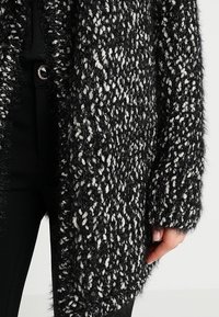 Czarny i biały dziergany kardigan o puszystej fakturze, z luźnym krojem i otwartym przodem, zestawiony z czarnymi spodniami.