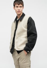 Béžový fleecový vest s černou džínsovou bundičkou pod ním, která má klasický límeček, kapsy a kontrastní textury. Nosit pod světlými cargo kalhotami.