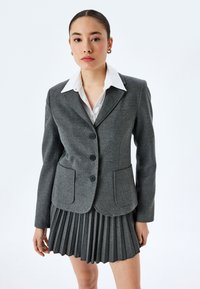 Grijze wollen blazer met een gelapte kraag, drie zwarte knopen en twee voorzakken, gecombineerd met een geplooide grijze rok.