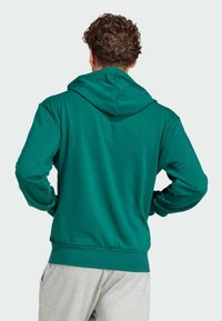 Zelená mikina s kapucňou na šnúrku, mäkká textúra a voľný strih. Môže obsahovať rebrované manžety a lem, viditeľné švy.