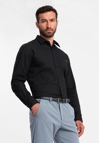 Zwarte overhemd met knopen, lange mouwen, kraag, borstzak. Gecombineerd met lichtgrijze broek en een zwarte riem. Glad, effen stoftextuur.