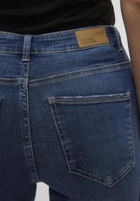 Tamno plave traperice s visokim strukom, kontrastnim šavovima i smešnim kožnim logom. Imaju dva zadnja džepa i glatku teksturu.