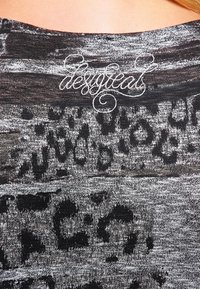Tessuto con stampa leopardata nera e grigia caratterizzato da una texture morbida, con "desygeat" ricamato in corsivo vicino al colletto.