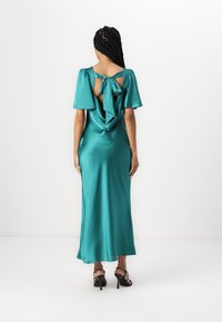 Abito maxi in satin teal con maniche a bufalo corte, schiena scoperta con dettagli a fiocco e un design drappeggiato. Indossato con tacchi neri a fascia.