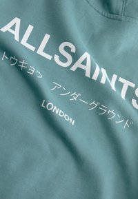 Türkizkék pamutszövet fehér felirattal. Tartalmazza az "ALLSAINTS" és a "LONDON" feliratokat japán nyelvű további szöveggel. Sima tapintású, lezser kialakítású.