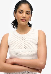 Ärmelloses Strickoberteil in Creme mit einem strukturierten Zickzackmuster und geripptem Ausschnitt. Das Model hat dunkles Haar, das nach hinten frisiert ist, und eine glatte Haut.