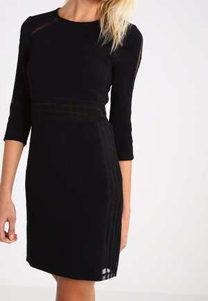 Kobieta ubrana w dopasowaną czarną sukienkę z rękawami do połowy przedramienia oraz teksturowanymi pionowymi i poziomymi paskami na jednolitym białym tle.