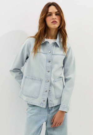 Vrouw draagt een lichtblauwe spijkerjasje met drukknoopsluiting en grote voorzakken, gecombineerd met bijpassende spijkerbroek, tegen een effen achtergrond.