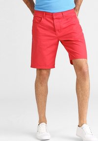 Red cotton shorts with a classic cut, featuring front pockets and a button closure. Worn with white sneakers.