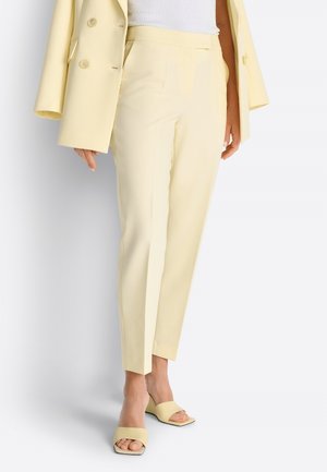 Donna che indossa pantaloni sartoriali giallo pallido, blazer abbinato posato sulle spalle, top bianco e sandali con tacco giallo pallido.