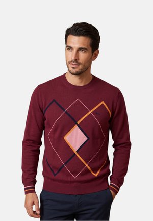 Uomo che indossa un maglione bordeaux con motivi geometrici a diamante in rosa, blu navy e arancione, abbinato a pantaloni blu scuro.