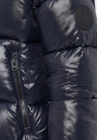 Marineblauwe pufferjack met glanzende textuur, voorzien van een ritszak op de linkermouw en een rond logo-embleem.