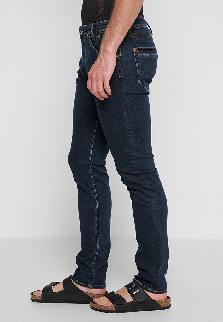Ein Mann steht seitlich, trägt dunkle Jeans und Sandalen und zeigt den Schnitt und Stil der Jeans vor einem neutralen Hintergrund.