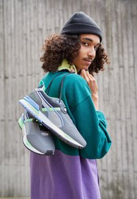 Gray sneakers with green and blue accents, mesh panels, and padded collars, hanging from a black strap over a green and purple outfit.