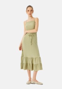 Abito midi in verde oliva, a coste, con spalline sottili, vita legata e orlo a strati con volant. Abbinato a scarpe slip-on chiare e decorate.
