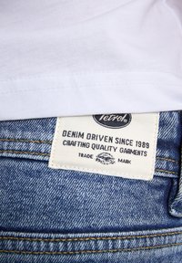 Vit bomull t-shirt över lager av blå denim jeans. Jeans har en tygetikett med svart text som säger "Denim Driven Since 1989."