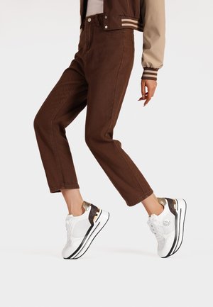Sneaker bianchi con mesh testurizzato e dettagli dorati, indossati con pantaloni ampi in corda marrone, su uno sfondo neutro.