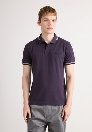 Young man wearing a dark purple polo shirt with striped collar and cuffs, paired with light grey pants, standing against a plain background.