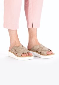 Beige textured slides with a cross-band design, featuring a white ridged sole and cork-like detail, set against light pink cropped pants.