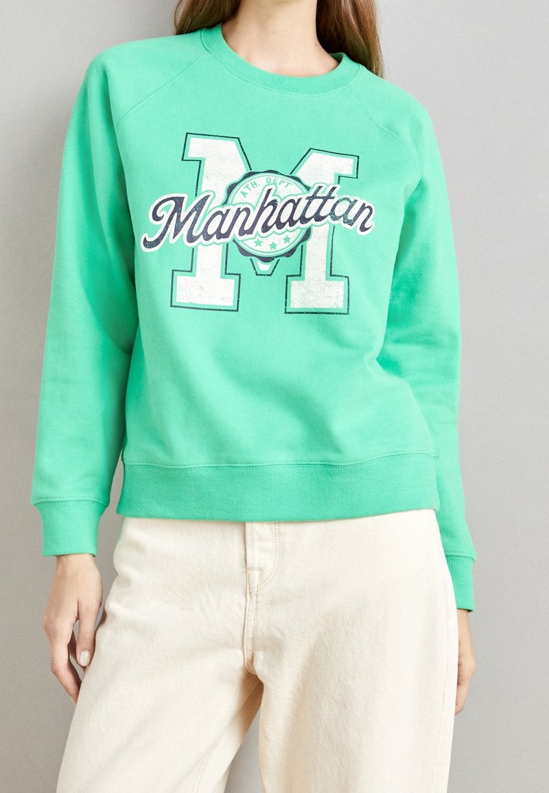 Persona vistiendo una sudadera verde menta con "Manhattan" y una gran "M" impresas en blanco y azul marino, combinada con pantalones beige claros.