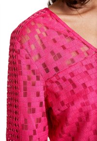 Blusa texturizada rosa con un patrón geométrico y escote en V. Presenta una tela semitranslúcida con acentos tejidos y detalles de punto abierto.