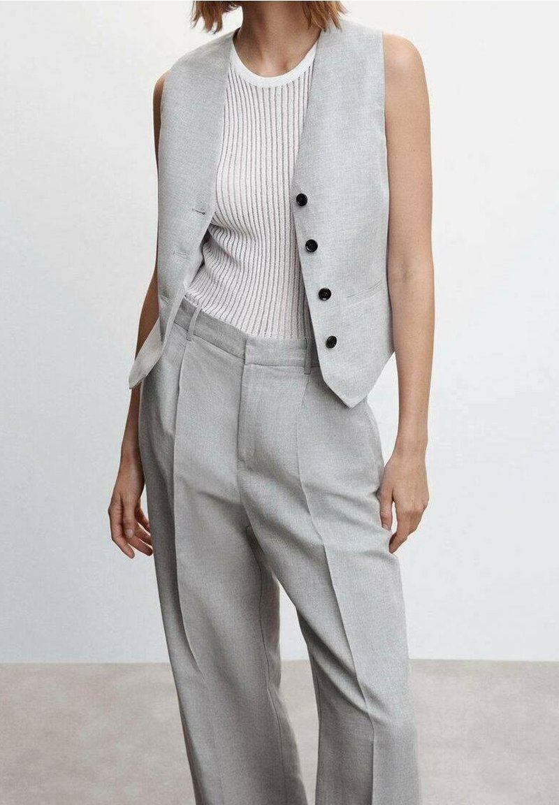 Personne portant un pantalon gris clair cintré et un gilet assorti par-dessus un top blanc côtelé sans manches, sur un fond uni.