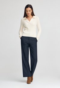 Cremfarbener, kabelgestrickter Pullover mit V-Ausschnitt und langen Ärmeln, kombiniert mit navyblauen, weiten Hosen und braunen Stiefeletten.