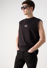 Juodas be rankovių marškinėliai su Calvin Klein logotipu, derinami su juodomis džinsomis. Modelis vilki stačiakampius akinius nuo saulės, demonstruodamas laisvalaikio stilių.