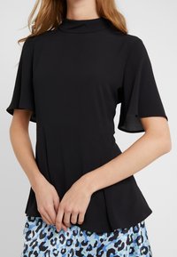 Czarna bluzka z wysokim kołnierzem i krótkimi, dzwonowymi rękawami; charakteryzuje się strukturalnym krój oraz gładką, elegancką fakturą. W zestawie z wzorzystą niebieską spódnicą.