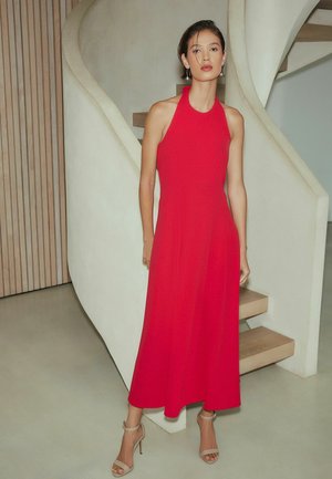 Žena stojaca v interiéri, oblečená v červenom midi šatách s odhalenými ramenami a béžových sandáloch na vysokom opätku, blízko modernej schodiska.