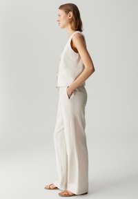 Beige mouwloos topje met een strikdetail aan de voorkant, gecombineerd met losse beige broeken. Platte sandalen maken de outfit af.