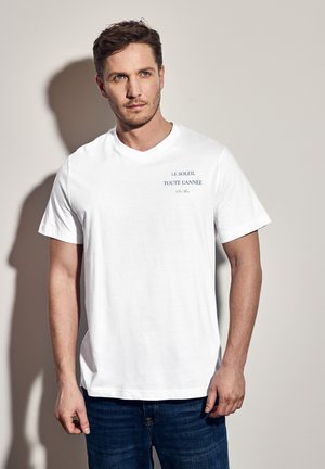 White cotton t-shirt with short sleeves, featuring blue text reading "LE SOLEIL TOUTE L'ANNEE." Simple, relaxed fit. Paired with blue jeans.