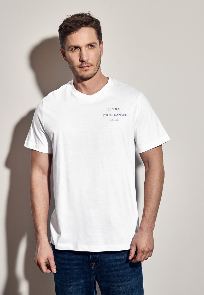 Valkoinen puuvillapaita lyhythihaisilla, jossa on sininen teksti "LE SOLEIL TOUTE L'ANNEE." Yksinkertainen, rento malli. Yhdistetty sinisiin farkkuihin.