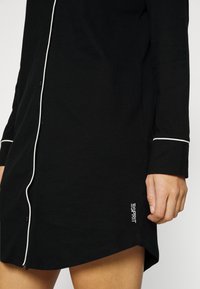 Camisa de noite preta feita de um tecido suave, com detalhes brancos ao longo da frente e das mangas, e um logótipo bordado na bainha.