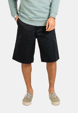 Hombre con pantalones cortos negros hasta la rodilla, zapatillas deportivas gris claro y sudadera verde claro, con las manos parcialmente en los bolsillos.