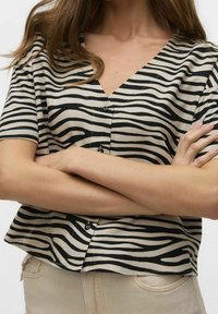 Haut à manches courtes beige avec des rayures zebra noires. Design en V avec des accents de boutons et une coupe décontractée et cropped. Texture de tissu douce.