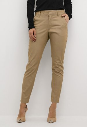 Osoba ubrana w beżowe wąskie spodnie z przednimi kieszeniami, zestawione z czarną prążkowaną bluzką z długim rękawem oraz beżowymi szpilkami w szpic z metalicznymi zakończeniami.