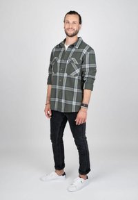 Groen geruite flanelhemd met twee borstzakken, gedragen over een wit T-shirt. Gecombineerd met zwarte jeans en witte sneakers. Effen achtergrond.