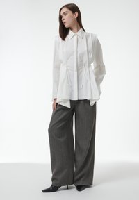 Camisa blanca de manga larga con cuello puntiagudo y detalle de cordón, combinada con pantalones anchos grises texturizados y tacones negros de punta.