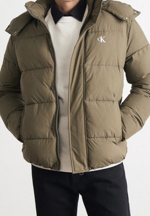 Homme portant une doudoune matelassée kaki à capuche par-dessus un pull blanc et une chemise noire à col, mains dans les poches, logo sur la poitrine.