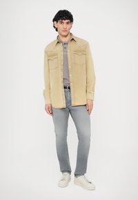 Chemise en velours côtelé beige clair avec deux poches poitrine, polo gris en dessous, jean gris clair et baskets blanches. Le modèle pose devant un fond blanc.