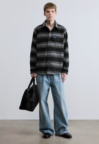 Μπλούζα από φανέλα με μαύρες και γκρι ρίγες και δύο τσέπες στο στήθος, σε συνδυασμό με φαρδιά ανοιχτό μπλε τζιν και μαύρη τσάντα duffel. Μαύρα επίπεδα παπούτσια.