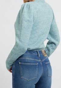 Sweter z dzianiny w jasnym turkusowym kolorze z bufiastymi rękawami, zestawiony z ciemnoniebieskimi dżinsami z metką na tylnej części pasa oraz tylnymi kieszeniami na łatę.
