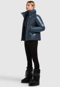 Jachetă albastră căptușită, cu guler înalt și textură lucioasă, combinată cu cizme negre impermeabile, purtate peste leggings negri.