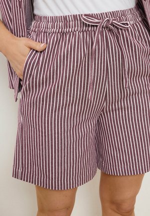 Hand in de linkerzak van paars-witte, verticaal gestreepte korte broek met trekkoord, gecombineerd met een bijpassend colbert en een wit shirt.