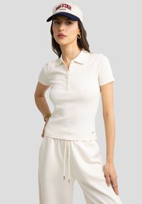 T-shirt polo en tissu ribbed crème avec un col plat, une patte de boutonnage à trois boutons et un branding discret. Associé à un pantalon blanc ample.