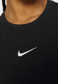 Detailní záběr osoby oblečené v černém žebrovaném topu s bílým logem Nike swoosh na hrudi.