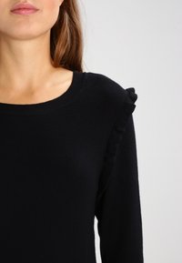 Svart tröja med rund halsringning och volangdetaljer vid axlarna. Tyget verkar mjukt och fint stickat.